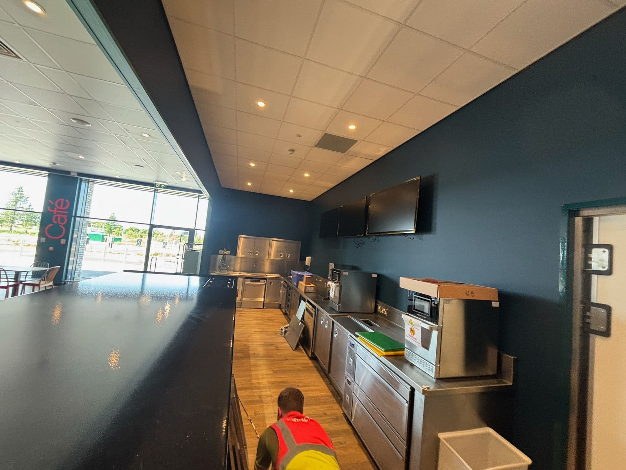 Long café servery with a dark feature wall and a team member in hi-vis working at the counter