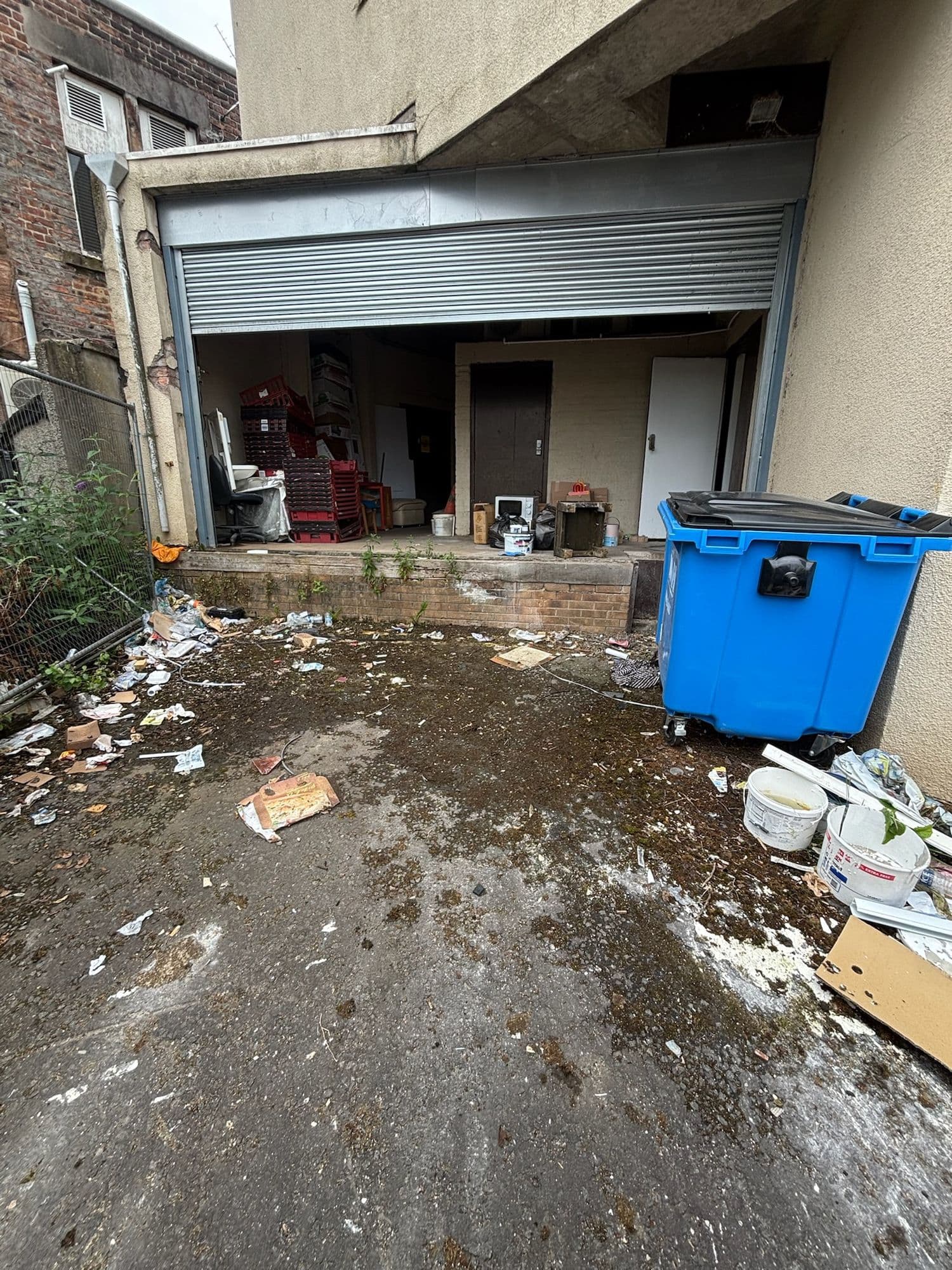Before: Rear service yard before cleaning, littered with rubbish and mud beside a blue commercial bin